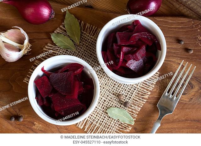 Fermented red beet with onions in two round bowls, with garlic, purple onions, allspice and bay leaf in the background, top view