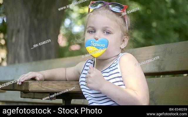Little blonde girl holds gingerbread in Ukrainian national colors in her hand, it says Ukraine is me. Close-up portrait of girl sitting on park bench with...