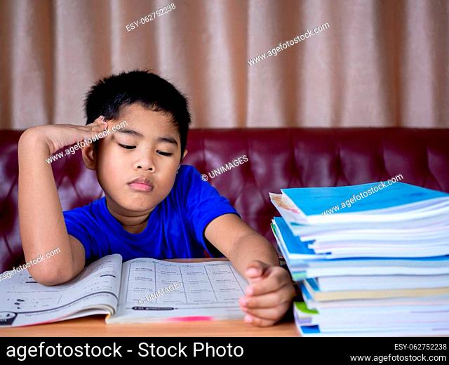 boy doing homework and reading on a wooden table with a pile of books beside The background is a red sofa and cream curtains
