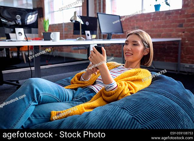 Happy mixed race businesswoman chilling in office relaxing space lying in beanbag using smartphone
