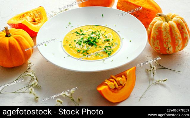 From above view of yellow pumpkin soup sprinkled with green herb and surrounded by pumpkins on white table