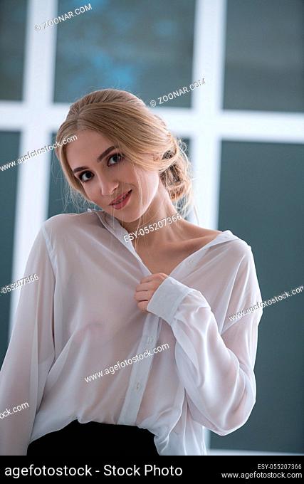 Beautiful woman wearing sexy white silk office shirt posing gracefully at camera in front of the window