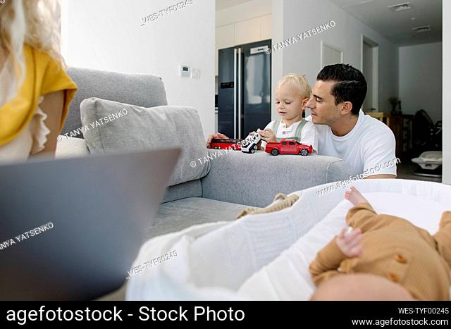 Man playing with son in front of baby in crib by woman working from home
