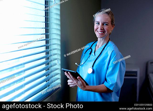 Smiling caucasian female doctor in hospital wearing scrubs and stethoscope holding tablet