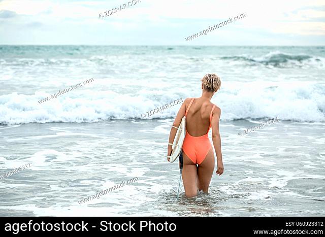 Blonde girl with short hairstyle stands backsides in the waves on the beach on the background of the sea and the cloudy sky