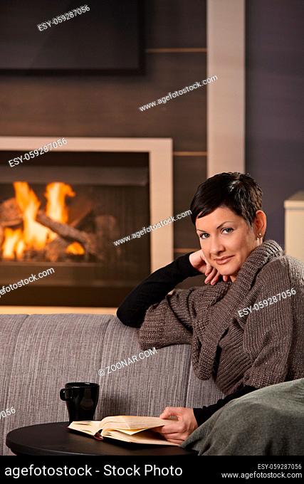Woman sitting on sofa at home on a cold winter day, reading book, looking up at camera