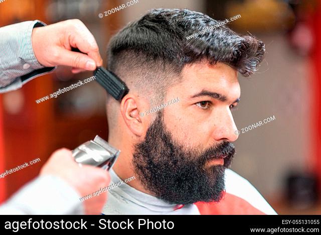 Handsome bearded man, getting haircut by barber, with electric trimmer at barbershop