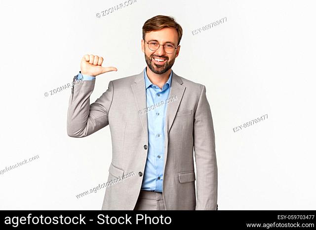 Portrait of confident and successful businessman with beard, wearing grey suit and glasses, pointing at himself and smiling, standing over white background