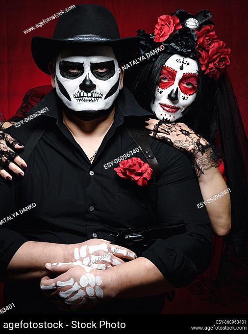 beautiful woman with a sugar skull makeup with a wreath of flowers on her head and a skeleton man in a black hat. Couple on dark red background