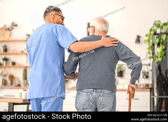 Back view of a male nurse supporting a gray-haired man leaning on the walking stick