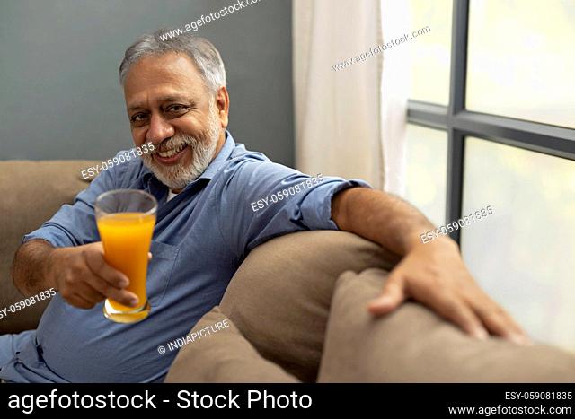 A SENIOR ADULT MAN SITTING ON SOFA AND OFFERING A GLASS OF FRUIT JUICE