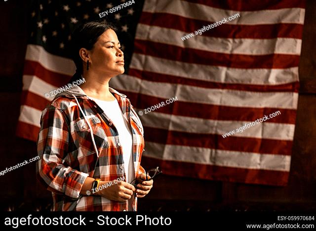 Patriotic Citizen Pondered Looking To Sideaway, A Beautiful Asian Woman In A Plaid Shirt Holding Glasses And Smiling On America's Flag Background