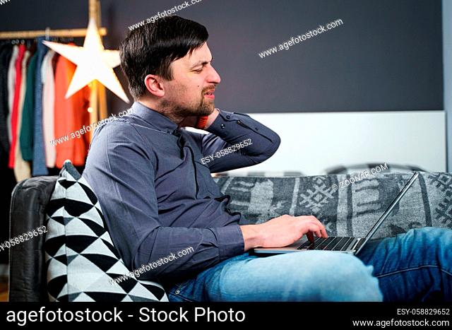 Tired strained man suffering from neck pain while working at computer. Male touchingly massages neck, puts on neck collar, sitting on couch