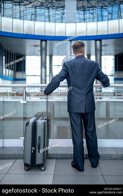 Wait. Man in elegant suit standing near suitcase touching railing with back to camera in terminal during day