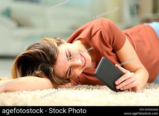 Happy woman reading message on mobile phone lying on a carpet on the floor at home