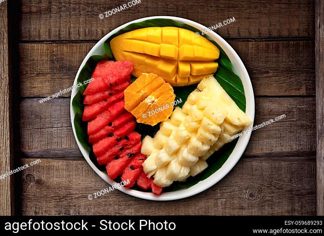 Top view fresh tropical fruits isolated on rustic wooden table. Mango, pineapple, watermelon and orange on a plate with banana leaf