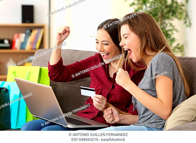 Two excited friends buying online with credit card and a laptop sitting on a sofa in the living room at home with a homey background