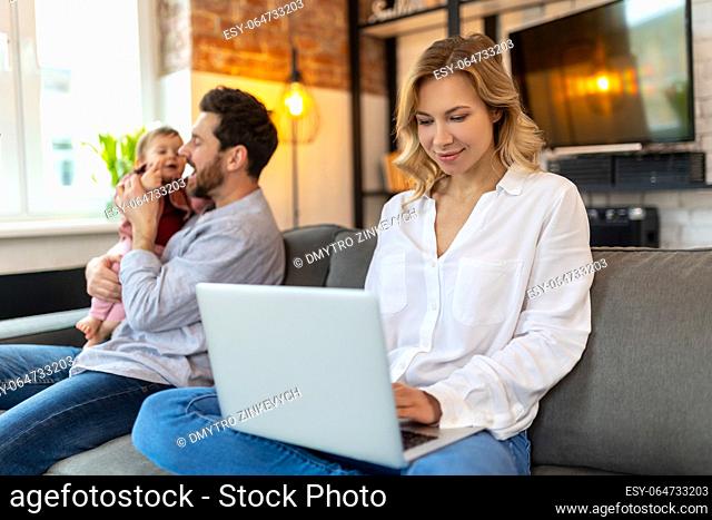 Father carrying infant daughter while mother using laptop on sofa remote work husband helping his wife with baby child