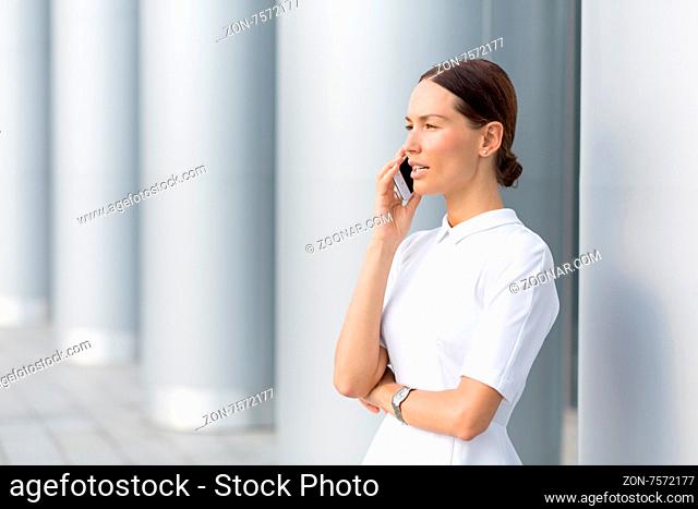 Confident young businesswoman keeping arms crossed and looking away while standing against white column. She is speaking over phone