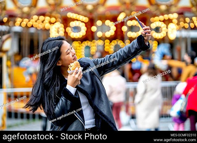 Woman holding burger and taking selfie through smart phone at park