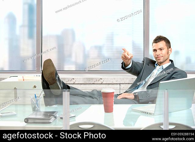 Relaxed businessman sitting at desk in front of office windows, pointing and looking at camera