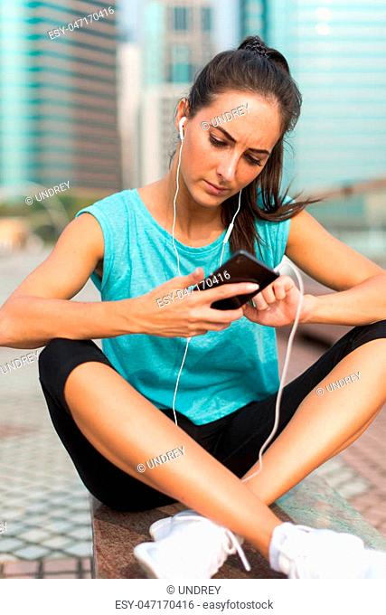 Young sporty woman resting after exercising using her cell phone and listening to music in earphones. Athlete runner in sportswear taking a break