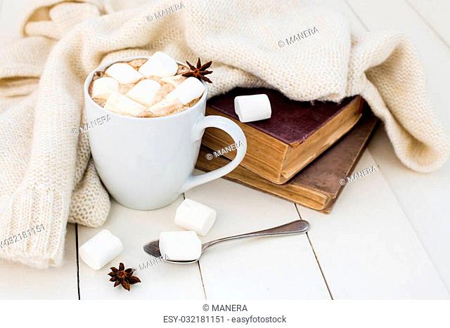 Cozy winter home background, cup of hot cocoa with marshmallow, old vintage books and warm knitted sweater on white painted wooden board background