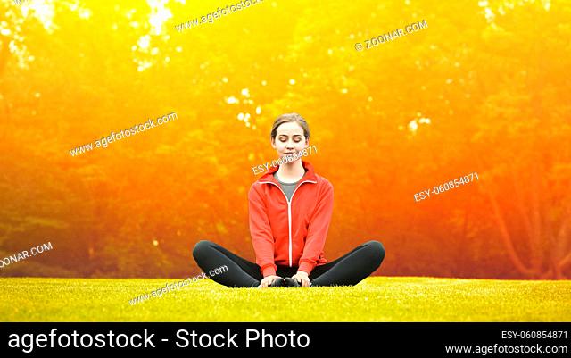 Pretty woman doing yoga exercises in green park or forest. Happy lady in red jacket sitting on green grass and looking at camera