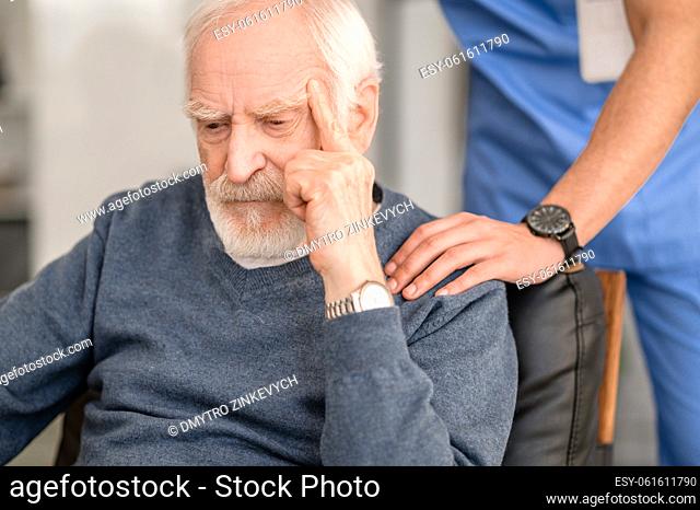 Sad old man sitting in the armchair while his caretaker patting him on the shoulder