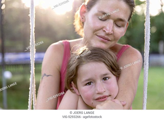 Mother sits in the sunshine on a swing in the garden and holds her daugther lovingly.- - Germany, 21/09/2010