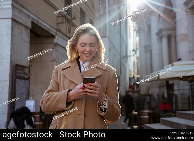 Smiling pleased blonde lady standing in the city center and looking at the smartphone screen