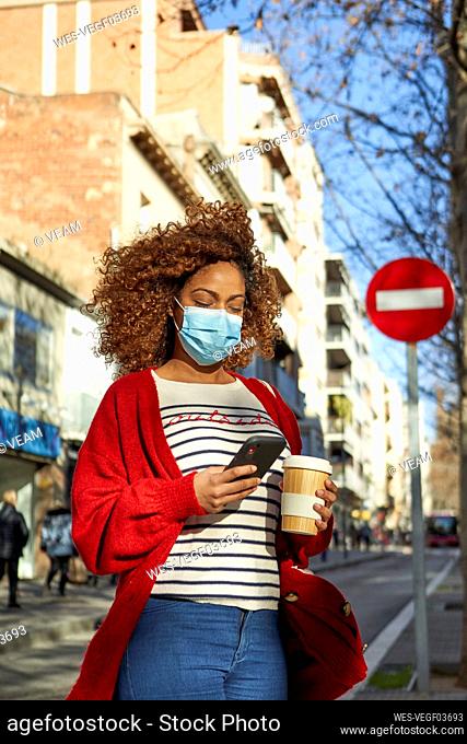Young woman wearing face mask using mobile phone while standing with coffee cup in city
