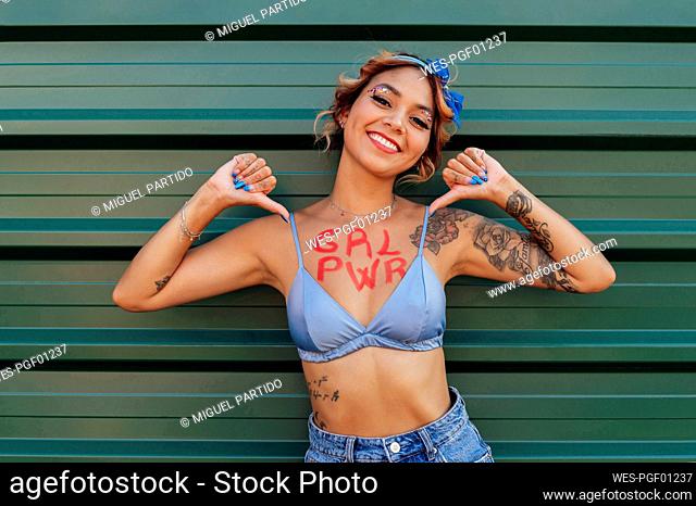 Happy young woman showing girl power text on chest in front of green wall