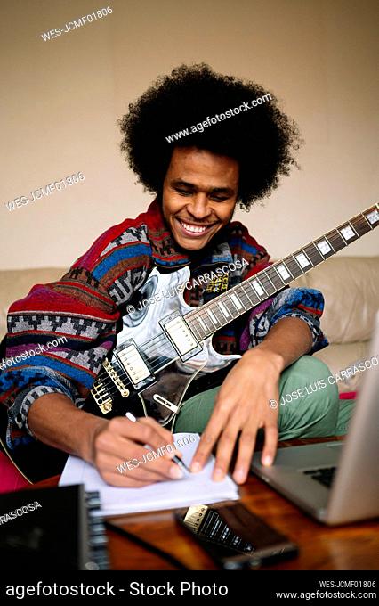 Happy male guitarist with guitar writing musical notes in book on table at home
