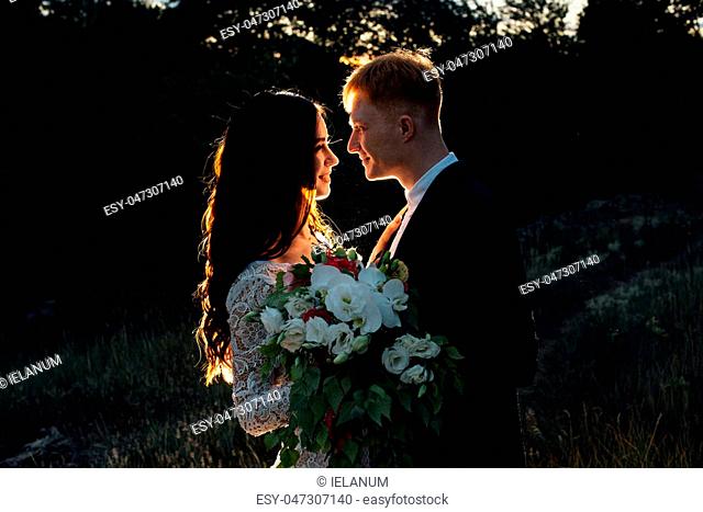 Newlyweds are standing and hugging outdoors, backlight, low key