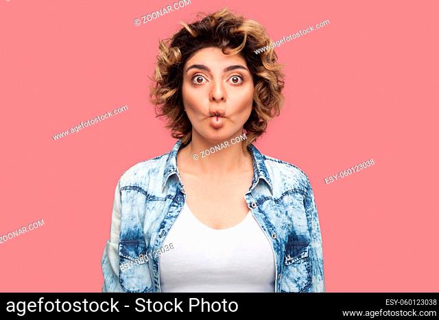 Portrait of funny young woman with curly hairstyle in casual blue shirt standing with fish pout lips and looking at camera with big eyes