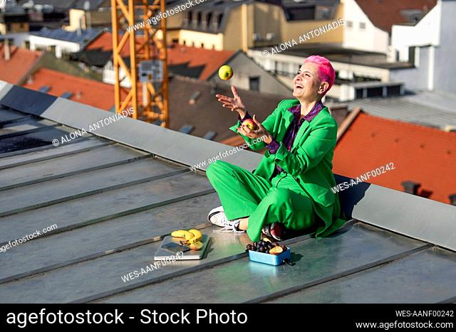 Cheerful businesswoman sitting cross-legged juggling apples on rooftop