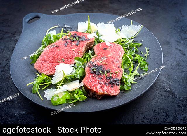 Modern style traditional fried New York strip steak with rucola and parmesan offered as close-up on a design plate