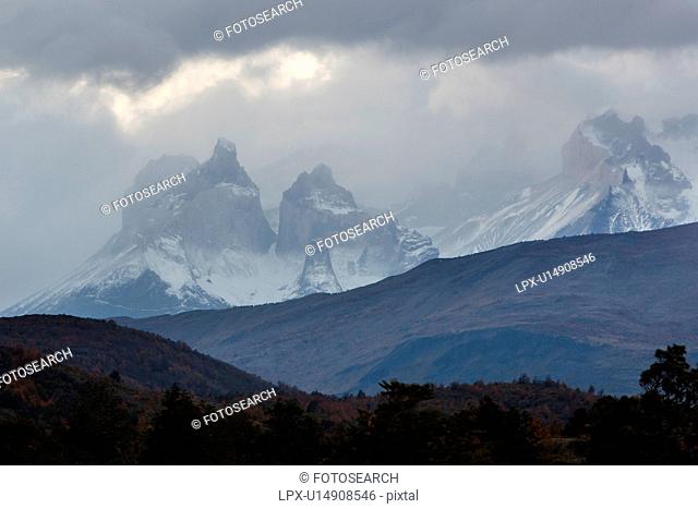 View of snow covered Los Cuernos mountains with threatening autumn sky, late afternoon, with hills and trees silhouetted in foreground, Torres del Paine