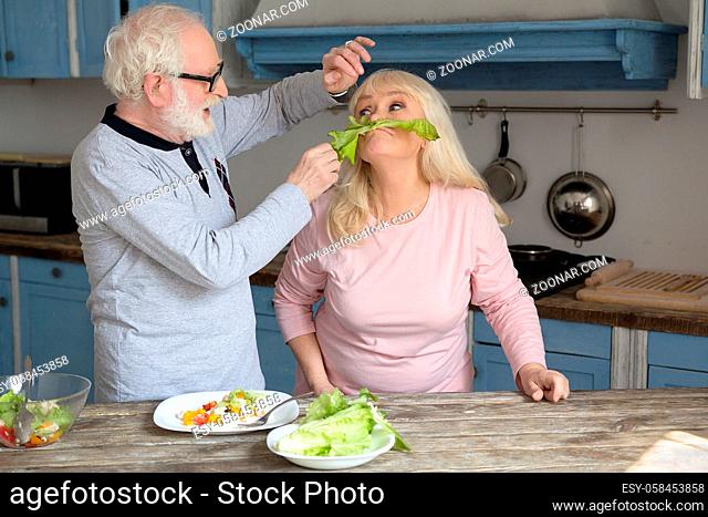 Old couple getting distracted while cooking. Two aged people wearing pajamas playing around with lettuce leaf in their kitchen during breakfast