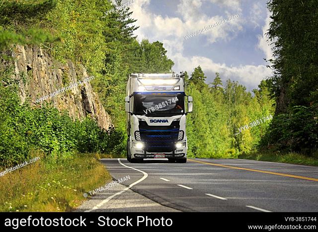 Customized Scania truck semi trailer Transport K. Lindholm & Co hauls load along highway, high beams on briefly. Salo, Finland. July 23, 2021