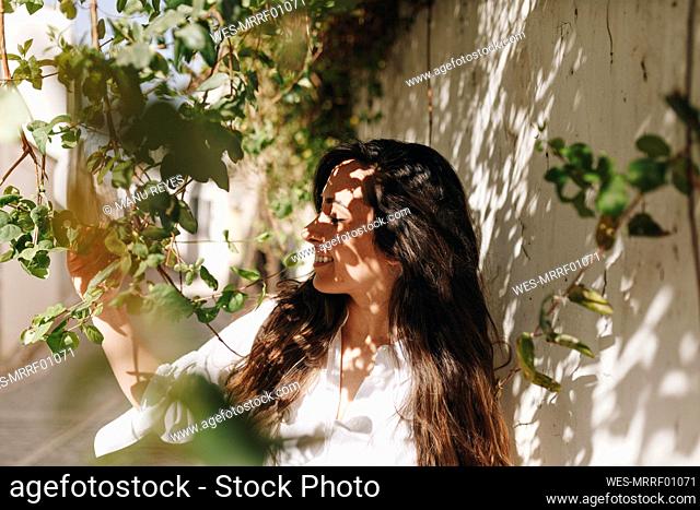 Woman smiling while touching plant by surrounding wall