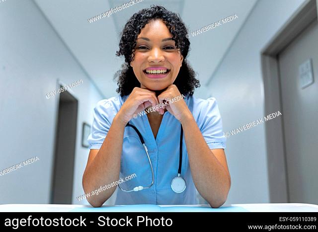 Mixed race female doctor at desk leaning chin on hands and smiling during video call consultation