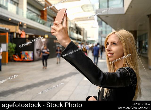 Close up portrait of an attractive young urban girl while taking selfie with her cell phone in the busy and crowded business district of London, United Kingdom