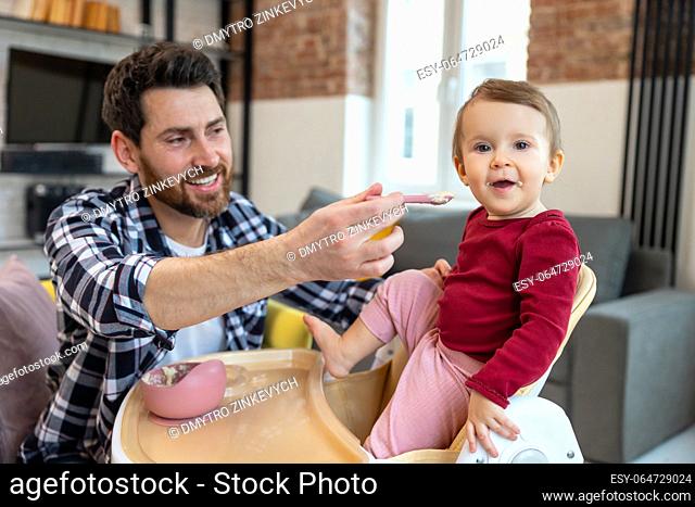 Lure first meal. Dad feeding toddler child with spoon for lunch in modern home interior