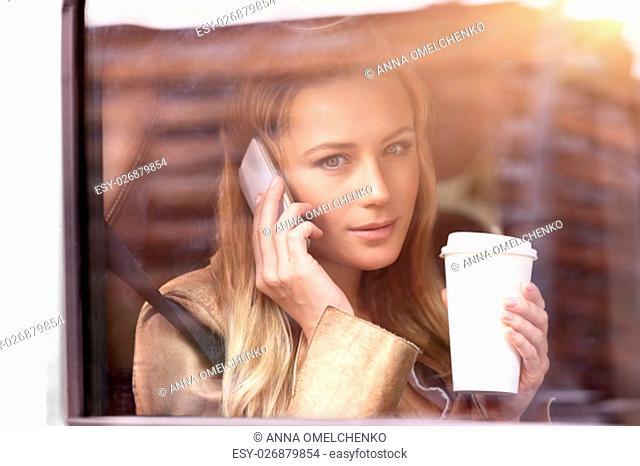 Portrait of a beautiful business woman sitting in the car with cup of coffee, talking on the phone and looking through car window, going to work