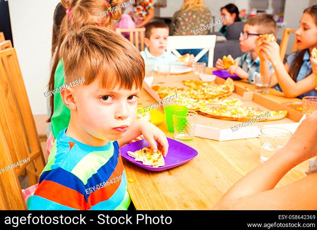 Children eating pizza at the wooden table, funny boy is looking at the camera