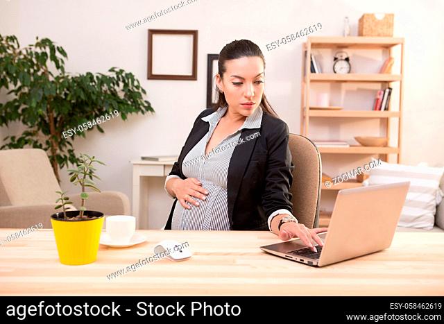 Young pregnant business woman working on laptop computer. Pretty lady touching her big belly while sitting at table. Business and freelance concepts