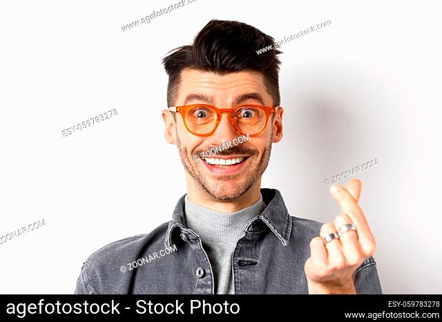 Close up portrait of cute guy with moustache wearing glasses, showing hand heart sign, smiling happy, standing on white background