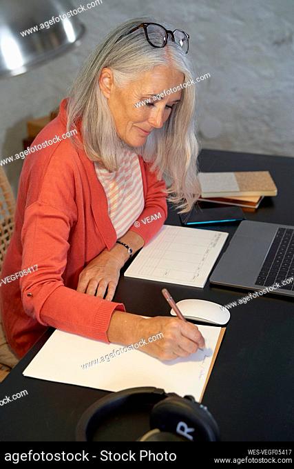 Smiling freelancer with gray hair writing on notepad at home